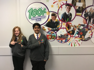 2 students stood in front of the “100% attendance club“ there are photos of other students on the wall, they are participating in various clubs, activities & rewards trips