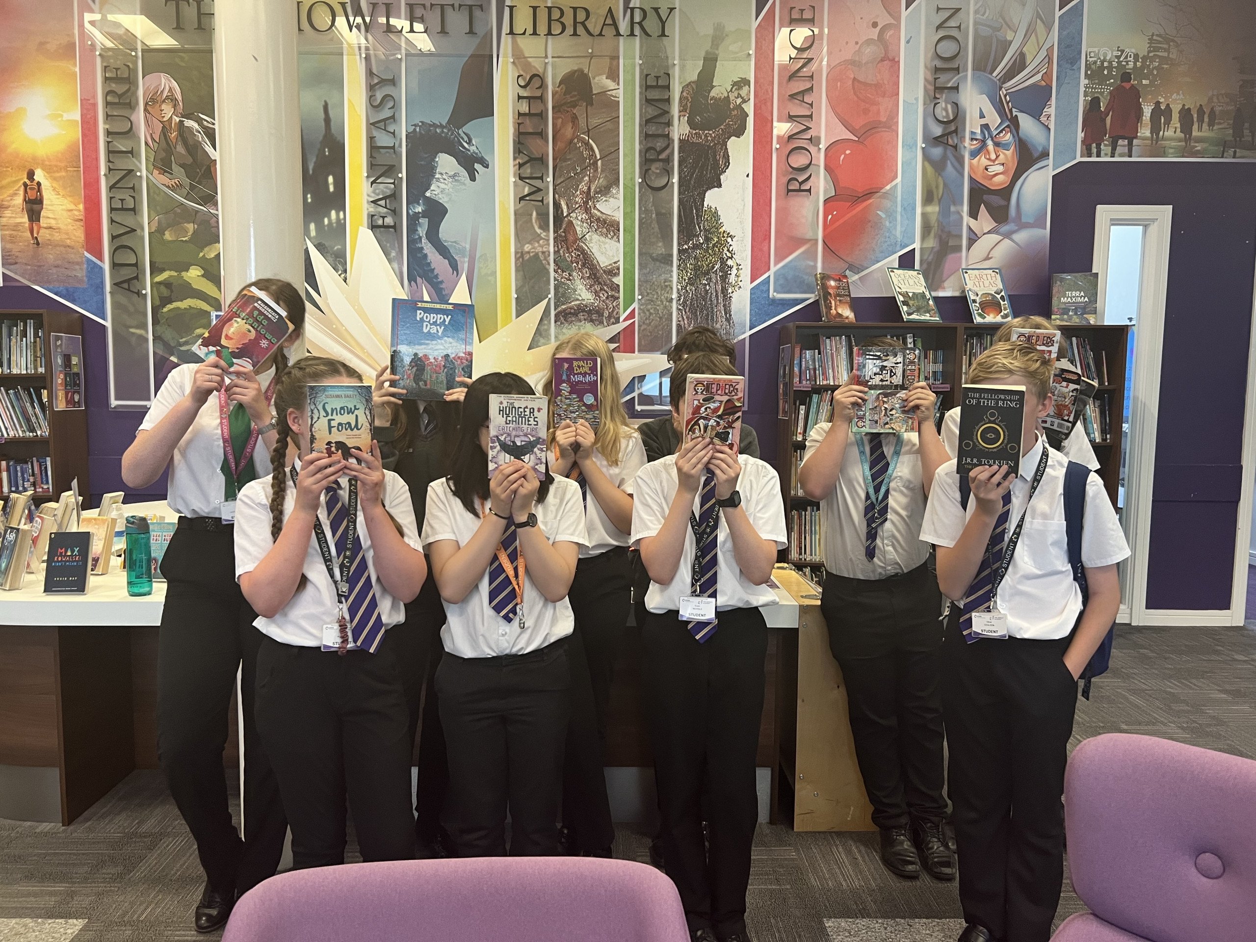 8 students stood in a library, they are all holding a book in front of their faces